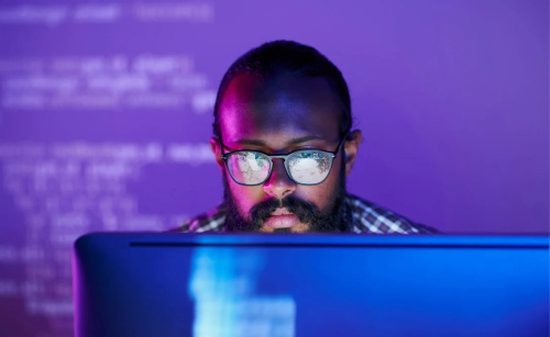 Bearded man with glasses working on a computer under purple and blue lighting