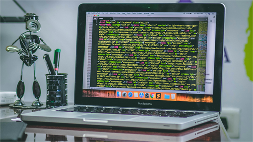 MacBook Pro laptop displaying colorful HTMLJS code on a desk with a robot figurine